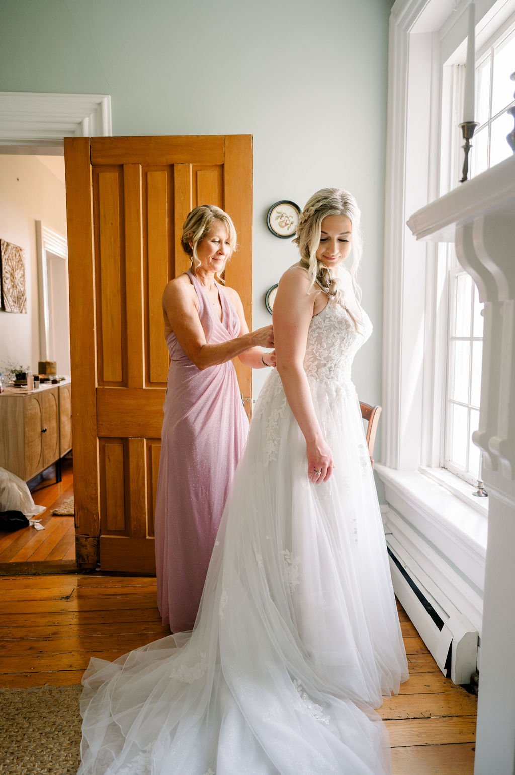 A bride in her gown beside a bright farmhouse window.