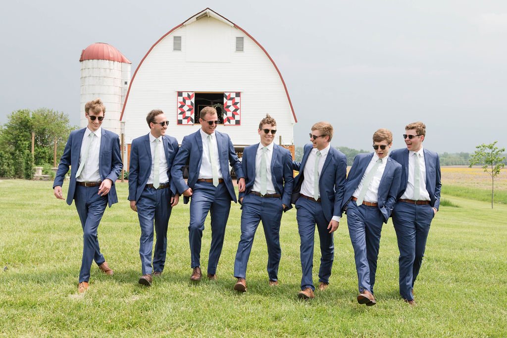 Seven groomsmen in blue suits walking across the field in front of the white barn and its quilt block.