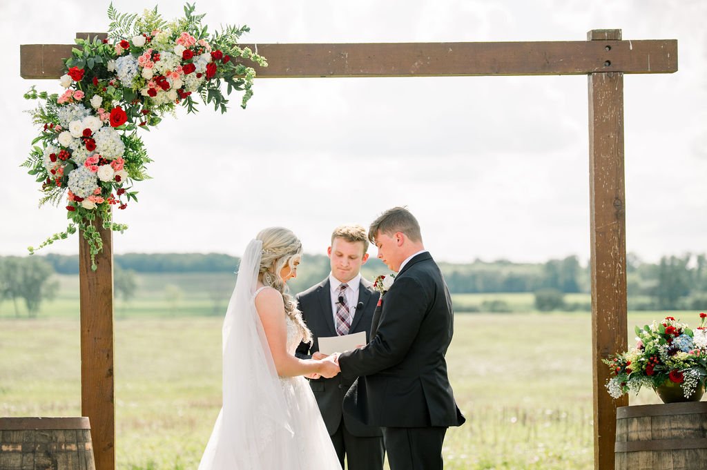 A couple stands at the flower-dressed outdoor arch in the open field.