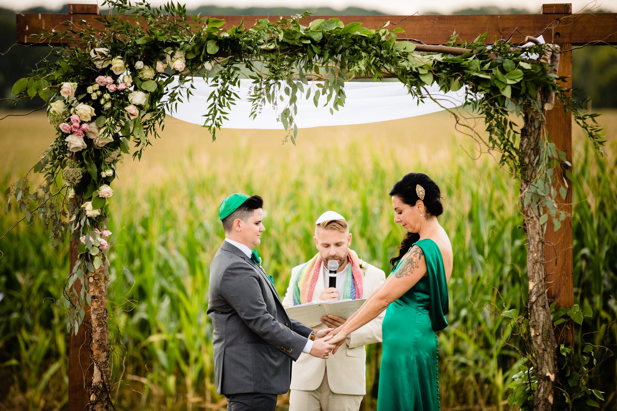 Two partners hold hands beneath the outdoor ceremony arch, cornfield behind them.