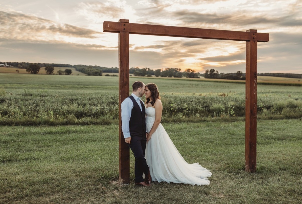 A couple framed by the wooden arbor out in the open field at sunset.