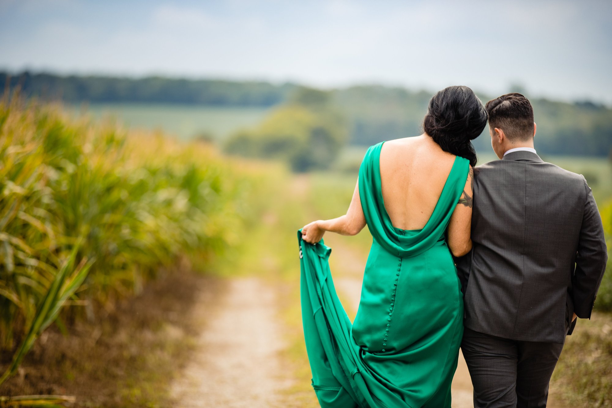 Two partners walk away down the farm lane between cornfields, one in an emerald gown.