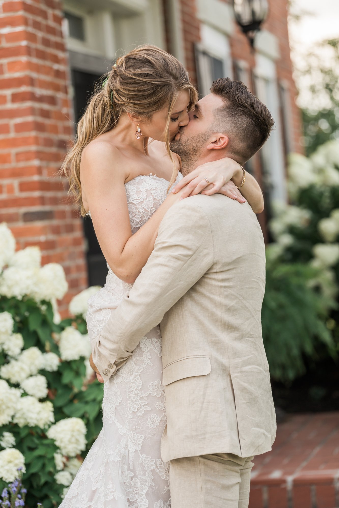 Groom lifts the bride for a kiss beside the brick farmhouse and white hydrangeas.