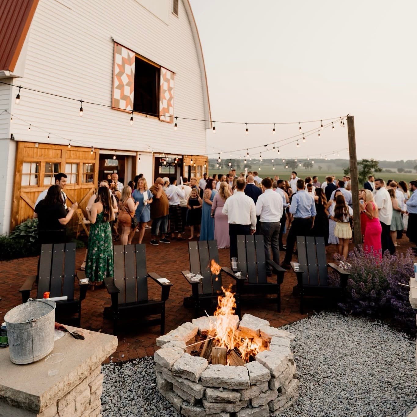 Cocktail hour on the brick patio at golden hour, guests under string lights with the fire pit lit.
