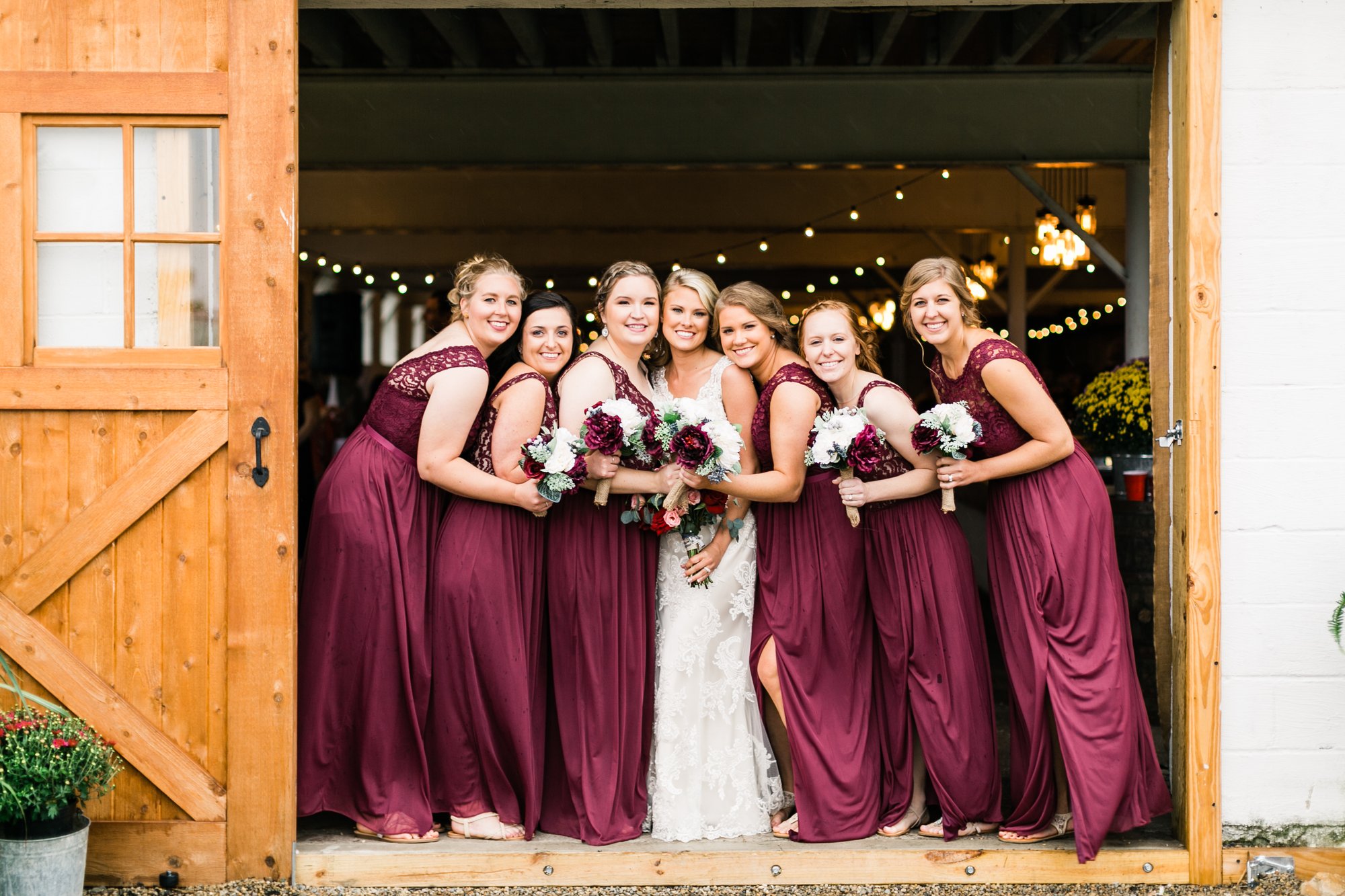 Bride and six bridesmaids in burgundy gathered at the open wooden barn doors.