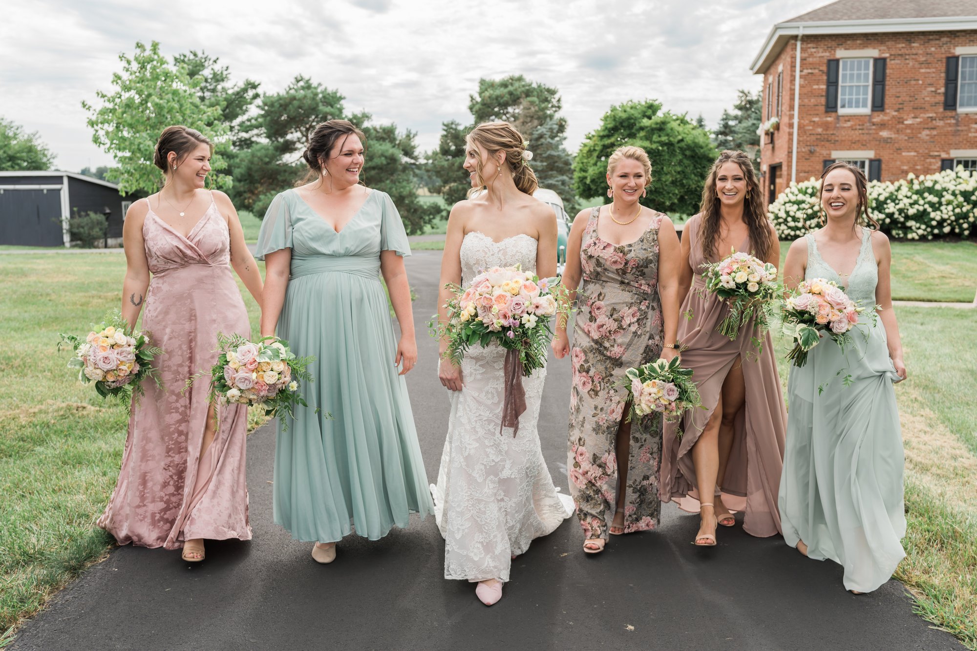 Bridesmaids in pastel gowns walking together past the brick farmhouse.
