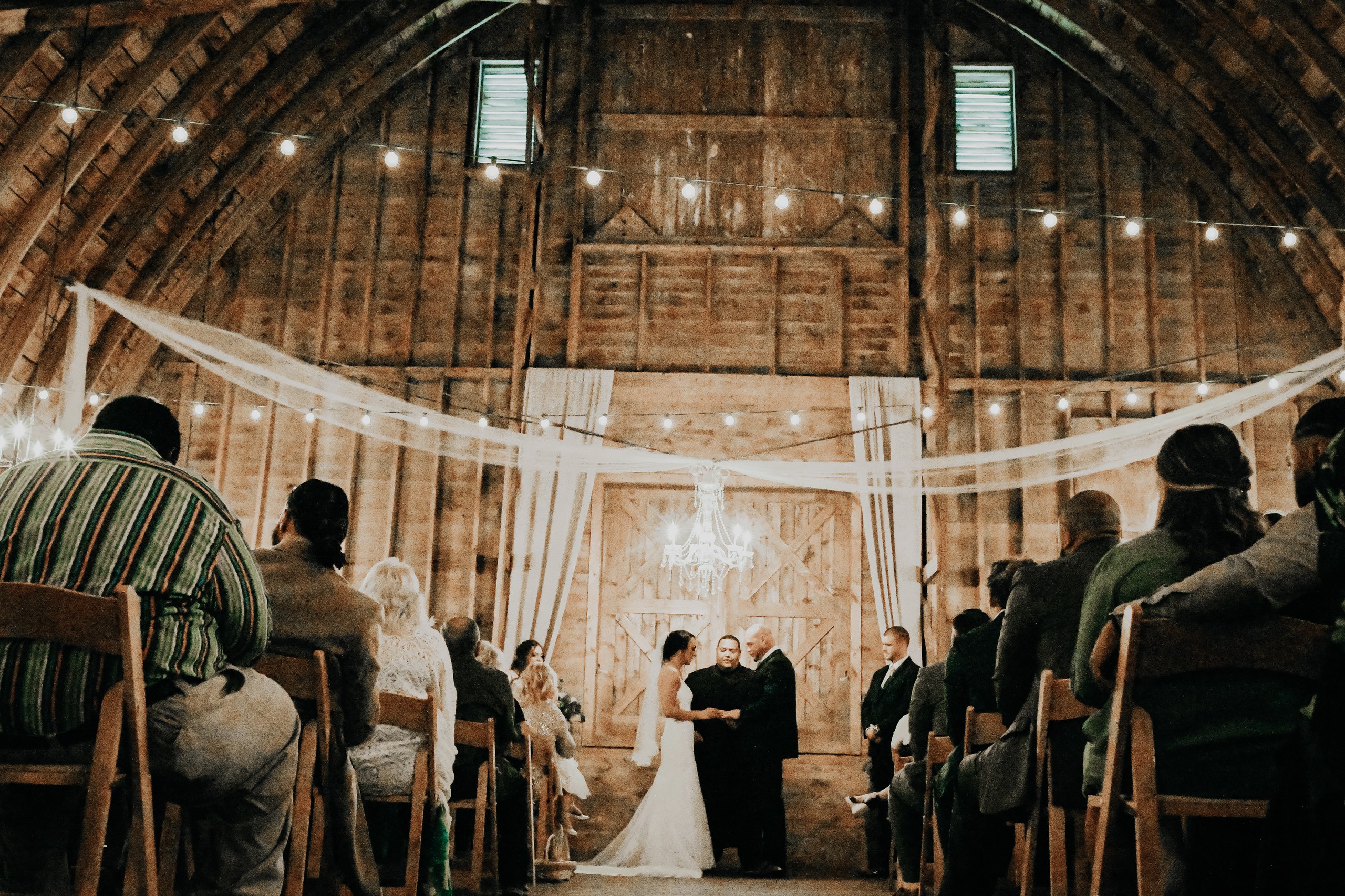 A wide view of an indoor barn ceremony with guests seated beneath the timber rafters.